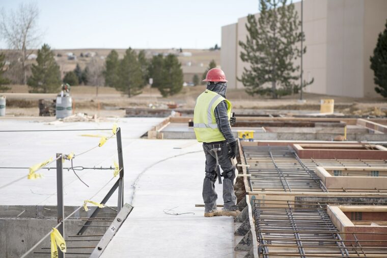 A person wearing a red hard hat and a yellow safety vest is standing at a construction site. There are trees in the background, and the person’s face is covered by a cloth mask.