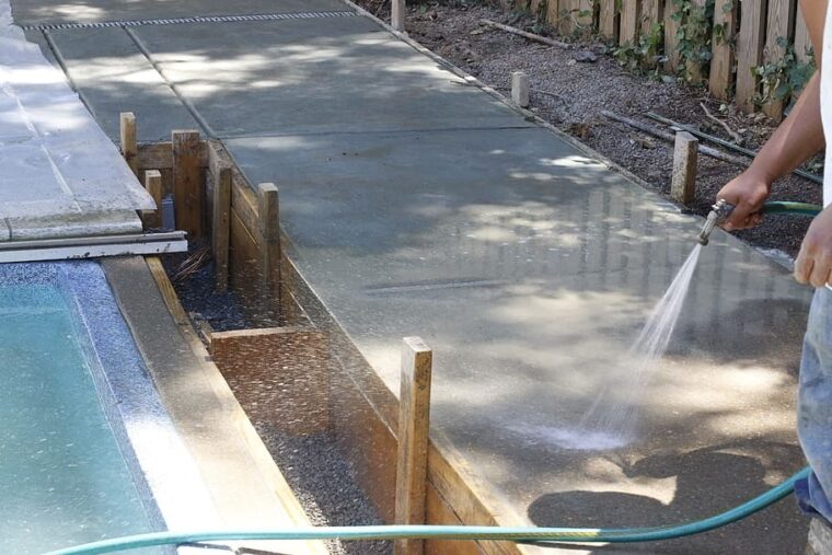 A person wearing blue jeans is spraying a concrete slab with a green water hose.