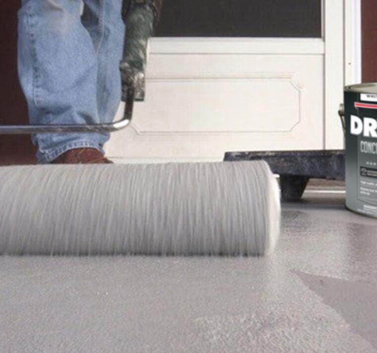 A person wearing blue jeans and brown shoes is applying grey, DRYLOK concrete paint on the floor.